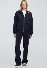 Chaqueta navy con cremallera y cuello en contraste rojo y blanco, combinada con pantalones de chándal navy y zapatillas blancas con detalles en marrón.