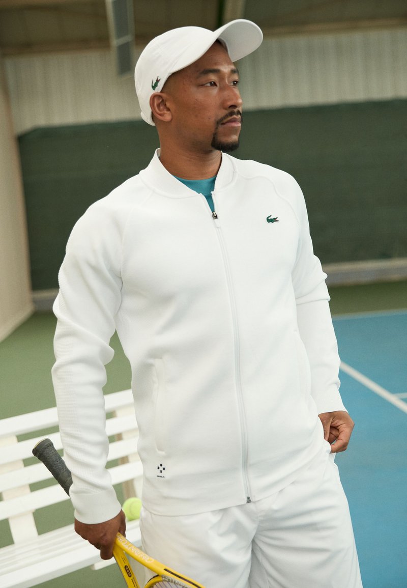 Veste blanche à fermeture éclair avec des poignets et un col côtelés, associée à des shorts blancs; tenant une raquette de tennis sur un court intérieur avec un fond vert.