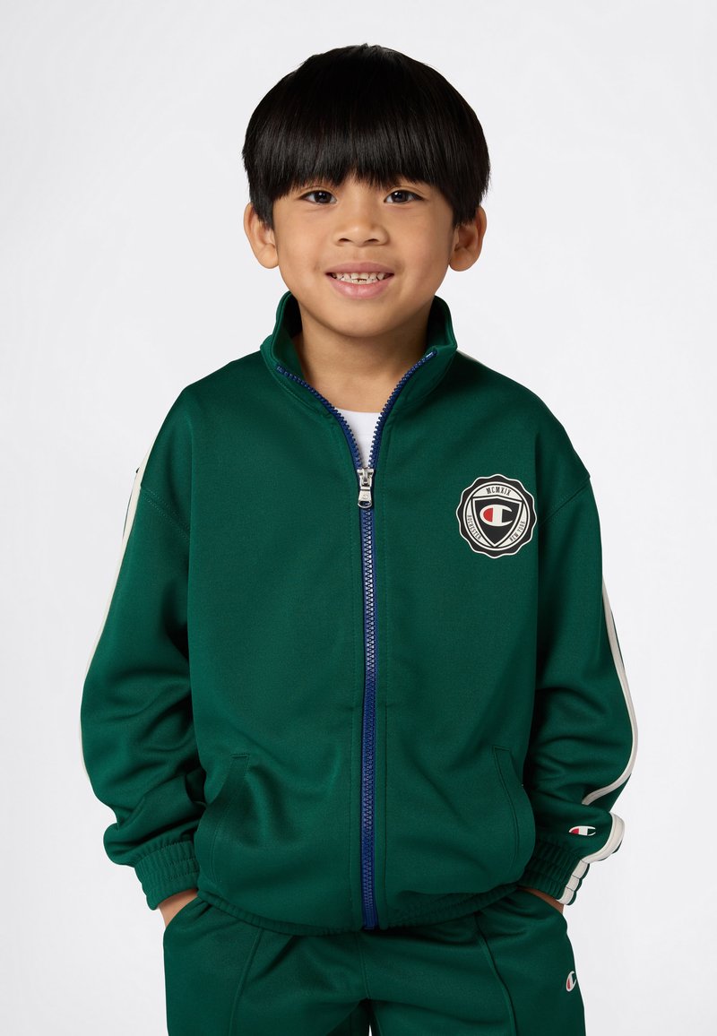 Niño pequeño sonriendo, vestido con un chándal verde con cremallera y franjas blancas y parches de logo en el pecho y la manga, con las manos en los bolsillos.
