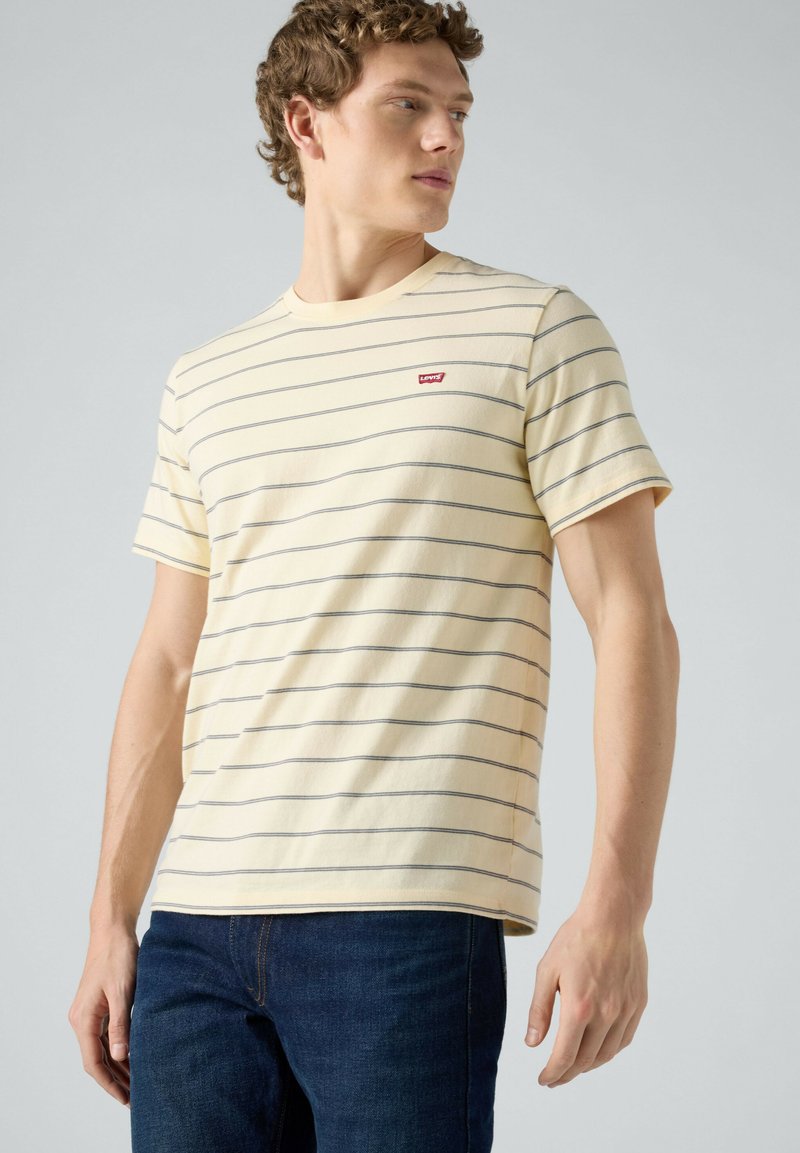 Jeune homme portant un T-shirt beige à fines rayures horizontales et un petit logo rouge Levi's, associé à un jean bleu foncé, regardant sur le côté.