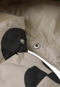 Beige vattentåligt tyg med stora svarta cirkulära mönster, med silverfärgade tryckknappar och synlig sömnad längs kanterna.