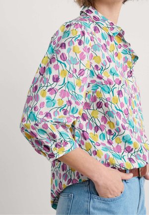 Femme portant un chemisier blanc à motif floral rose, jaune et bleu avec des manches bouffantes, rentré dans un jean bleu clair avec une ceinture marron.