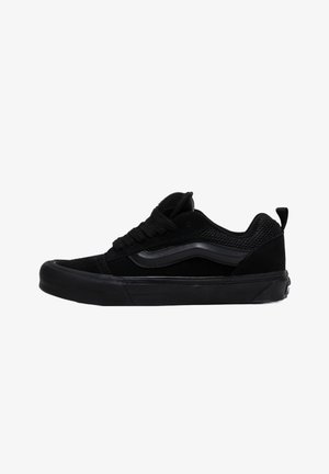 Black skate shoes made of suede and mesh, featuring a textured toe cap, flat laces, and a signature wave design on the side.