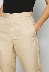Beige cotton trousers with a smooth texture, featuring a front button closure, side pockets, and relaxed fit. No visible patterns.