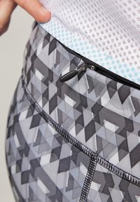 Leggings met een geometrisch grijs patroon en zwarte accenten, een ritssluiting op de tailleband en een gestructureerde blauwe mesh voering aan de bovenkant.