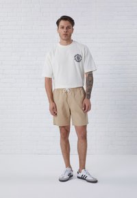 T-shirt bianca a maniche corte con logo grafico nero, abbinata a pantaloni corti beige con vita elastica e cordino. Sneakers grigie e nere.
