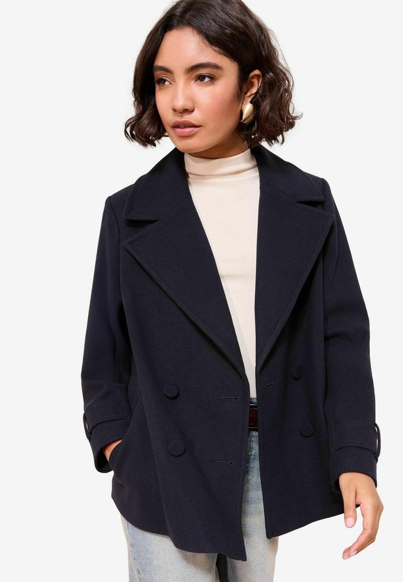 Femme aux cheveux courts et foncés portant des boucles d'oreilles en or, un manteau noir croisé, un pull col roulé beige et un jean bleu clair, les mains dans les poches.