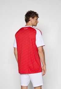 Rode en witte sporttrui met een gestructureerde rode achterkant met een patroon, korte mouwen en een ronde hals. Logo op de achterkant.