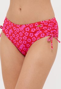 Nő piros bikini alsót visel rózsaszín virágmintával és a „SUN” felirattal, állítható oldalsó megkötőkkel és apró arany márkacímkével.