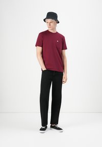 Bordeaux crew neck t-shirt med lomme, sorte bukser og sort bucket hat. Modellen har sorte snørebåndssneakers på og står foran en ensfarvet baggrund.