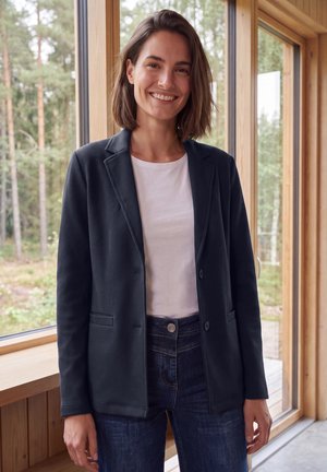 Femme aux cheveux bruns mi-longs souriante, portant un blazer foncé, une chemise blanche et un jean, debout dans une pièce en bois avec de grandes fenêtres montrant des arbres à l'extérieur.