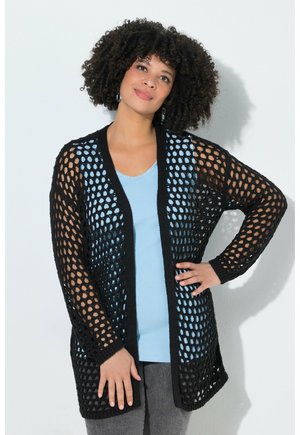 Kvinde med krøllet hår iført en sort åbenstrikket cardigan over en lyseblå top og grå bukser, stående med den ene hånd på hoften.