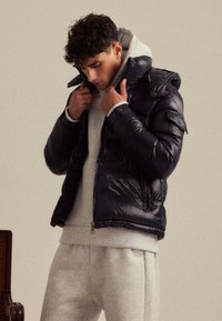 Doudoune matelassée bleu marine foncé avec une finition brillante, dotée d'une capuche, d'une fermeture éclair et d'accents de poche. Portée par-dessus un sweat à capuche gris clair et un bas assorti.