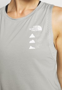 Osoba oblečená v světle šedém bezrukávovém tričku North Face s černou vnitřní vrstvou, na hrudi s horskými grafikami a vytištěnými roky.