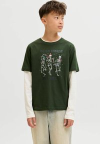 Garçon aux cheveux courts portant une chemise à manches longues verte et blanche à motifs de squelettes et avec le texte "Mort à l'intérieur mais joyeux", regardant vers la droite.