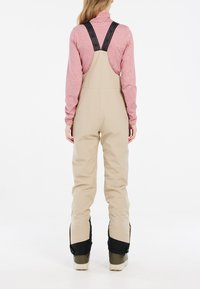 Beige waterdichte overall met verstelbare zwarte banden, taps toelopende benen en een duurzame textuur. Kenmerken zijn onder meer verstevigde zwarte enkelpanelen.