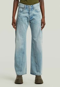 Persoon in lichtblauwe wijde jeans en olijfgroene platformlaarzen, staande tegen een effen lichte achtergrond.