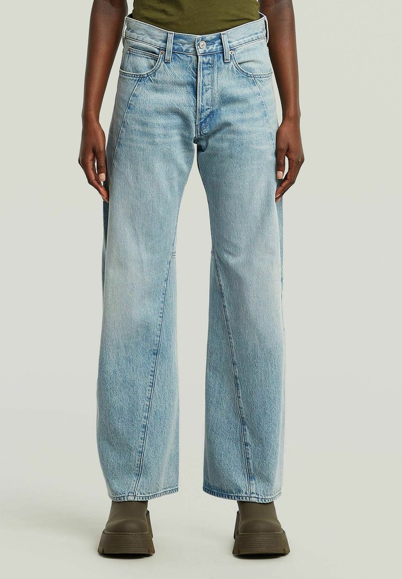 Persoon in lichtblauwe wijde jeans en olijfgroene platformlaarzen, staande tegen een effen lichte achtergrond.