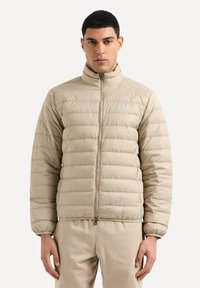 Giacca puffer beige con chiusura a zip, colletto alto, design trapuntato e piccolo logo sul lato sinistro del petto. Materiale liscio e leggero.