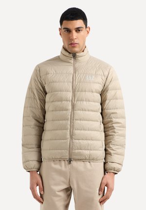 Giacca puffer beige con chiusura a zip, colletto alto, design trapuntato e piccolo logo sul lato sinistro del petto. Materiale liscio e leggero.