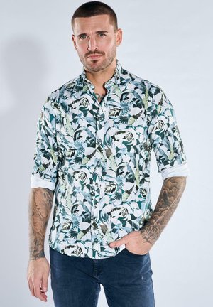 Mann mit Tätowierungen, der ein hochgekrempeltes, geblümtes Hemd mit Knöpfen und dunkle Jeans trägt, mit der Hand in der Tasche vor einem einfarbigen Hintergrund steht.