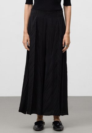 Zwarte geplooide broek met wijde pijpen en een elastische tailleband, voorzien van verticale strepen en een lichte stoffen textuur. Gepaard met zwarte instapschoenen.