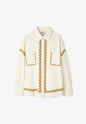Camisa crema de manga larga con botones, cuello puntiagudo y ribete marrón decorativo bordado en los bolsillos, el frente, las mangas y el dobladillo.