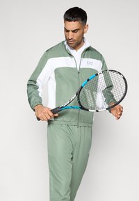 Πράσινο και λευκό αθλητικό μπουφάν με φερμουάρ, σετ με αντίστοιχο παντελόνι. Το μοντέλο κρατάει ρακέτα τένις με μαύρο και μπλε σχέδιο.