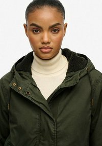 Veste à capuche vert foncé avec un col en polaire et un cordon de serrage. Superposée sur un col roulé crème. Dotée de quincaillerie dorée et de poches.
