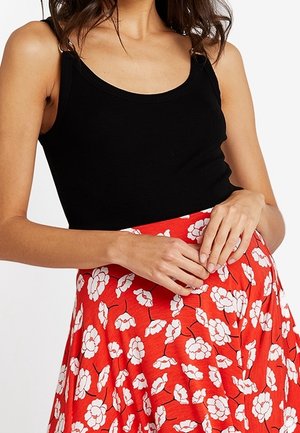 Femme portant un haut noir sans manches et une jupe rouge à imprimé floral blanc, les mains reposant doucement sur la taille de la jupe.