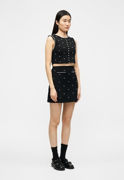 Γυναίκα με μαύρο crop top χωρίς μανίκια και ταιριαστό μίνι φούστα με διακοσμητικά μαργαριταριών, μαύρες κά λτσες και μαύρα loafers, στέκεται μπροστά σε λευκό φόντο.