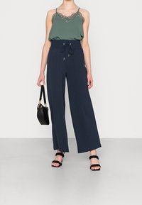 Camisole verte avec bordure en dentelle, associée à un pantalon large bleu marine avec taille à cordon. Sac à main noir et sandales à lanières complètent le look.