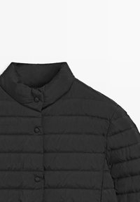 Veste matelassée noire avec col montant et trois boutons pression visibles sur un fond blanc.