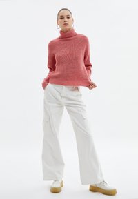 Pull en maille rose avec col roulé et une finition texturée, associé à un pantalon cargo blanc à jambes larges. Les chaussures présentent une semelle épaisse.