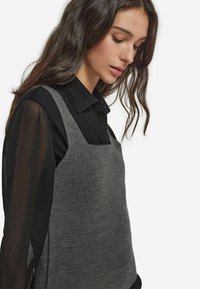 Szary dzianinowy top na ramiączkach z kwadratowym dekoltem, noszony na czarnej przezroczystej koszuli z długim rękawem. Gładka faktura.