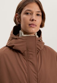 Bruine geïsoleerde jas met een hoge kraag, textielstof en een rits aan de voorkant. Heeft verstelbare hardware voor een betere pasvorm. Eenvoudig ontwerp.