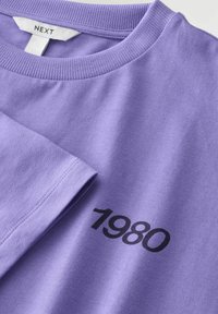 Lila pamučna majica s okruglim izrezom, kratkim rukavima i crnim otisnutim detaljem "1980" sprijeda. Glatka tekstura, opušteni kroj.
