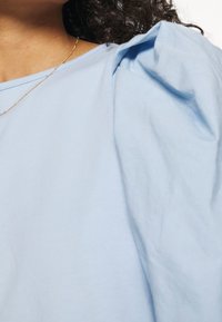 Top azul claro con mangas abullonadas, hecho de suave tela de algodón. El escote es redondeado y sutilmente acanalado.