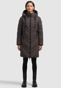 Donna che indossa un lungo cappotto puffa trapuntato marrone scuro con cappuccio, leggings neri e stivaletti neri, in piedi su uno sfondo semplice.