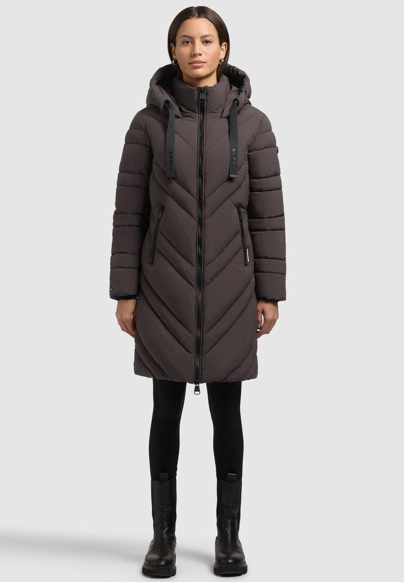 Donna che indossa un lungo cappotto puffa trapuntato marrone scuro con cappuccio, leggings neri e stivaletti neri, in piedi su uno sfondo semplice.