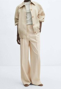 Beige oversized jacket with button front and raglan sleeves, paired with wide-leg trousers in a lighter beige. Smooth fabric and relaxed fit.