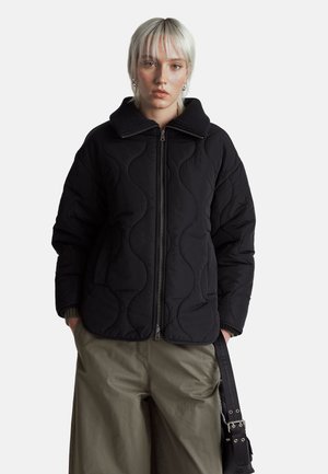 Czarna pikowana kurtka z wysokim kołnierzem, z falistym wzorem i zapięciem na zamek. Połączona z luźnymi zielonymi spodniami i czarnym paskiem.