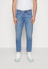 Calças de ganga azul claro com corte reto, apresentando cinco bolsos e fecho de zip, usadas com sapatilhas brancas, exibindo um design casual.