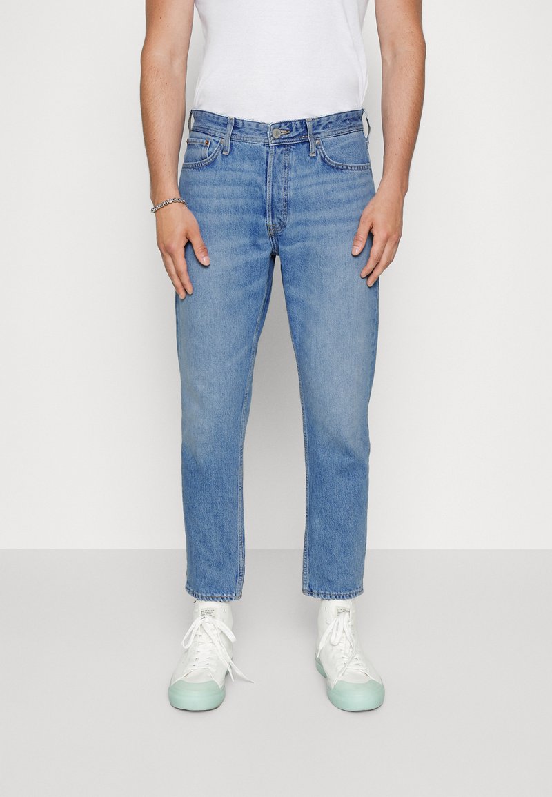 Calças de ganga azul claro com corte reto, apresentando cinco bolsos e fecho de zip, usadas com sapatilhas brancas, exibindo um design casual.