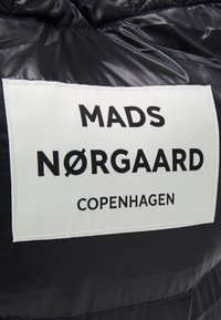 Zwarte pufferjack met een wit label waarop "MADS NØRGAARD" in vetgedrukte zwarte letters staat en daaronder "COPENHAGEN".