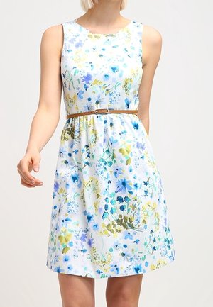 Woman wearing sleeveless white dress with blue, green, and yellow floral pattern and a thin brown belt at the waist.