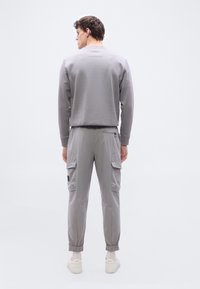 Άνδρας που στέκεται με την πλάτη, φοράει ανοιχτό γκρι φούτερ, γκρι παντελόνι cargo με τσέπες, λευκά καλτσάκια και λευκά αθλητικά παπούτσια σε λευκό φόντο.