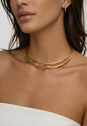 Donna che indossa una doppia collana a girocollo in oro e piccoli orecchini a cerchio in oro, con pelle liscia e un top bianco senza spalline.