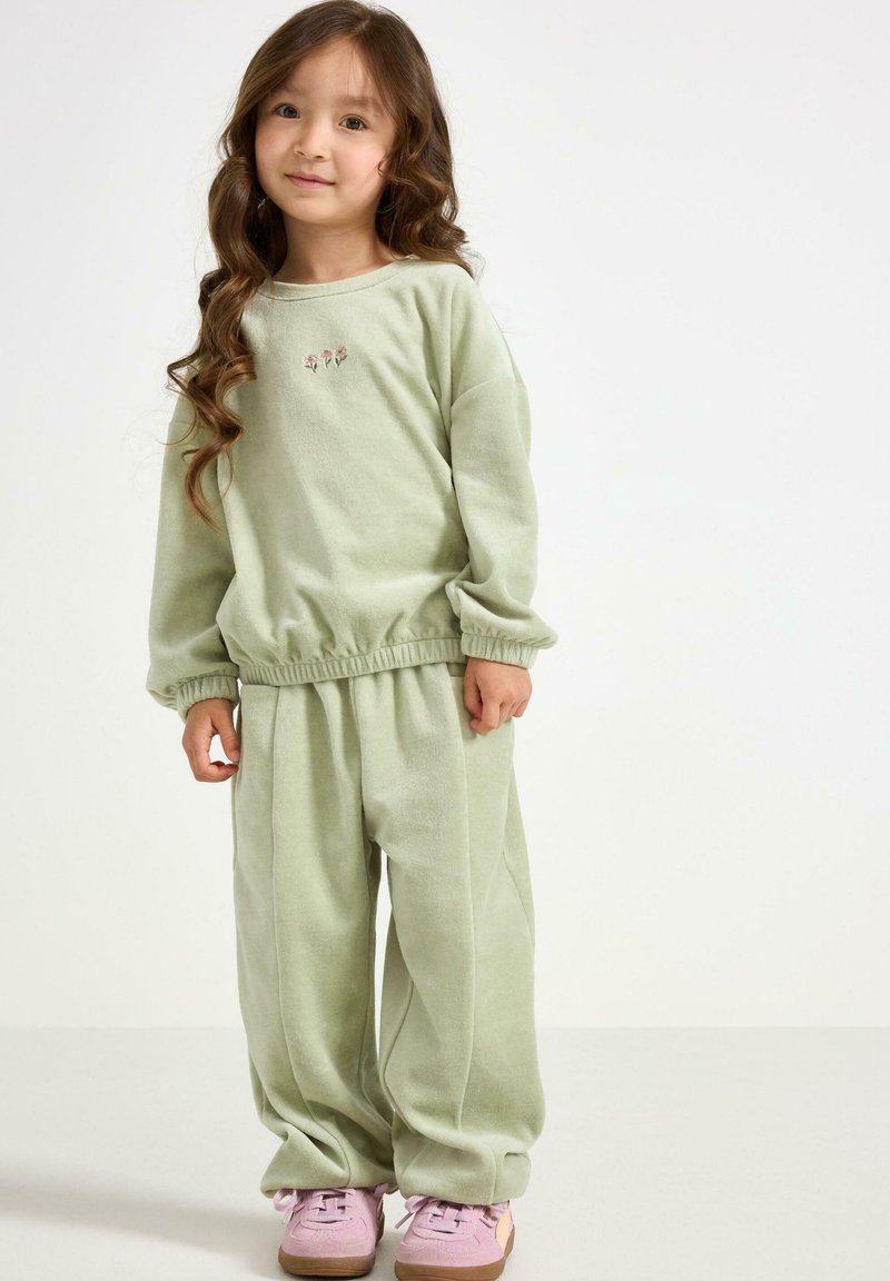 Grüner flauschiger Baumwoll-Trainingsanzug mit elastischen Bündchen und einem elastischen Bund, der florale Stickereien auf dem Oberteil aufweist. Kombiniert mit pinken Sneakers.
