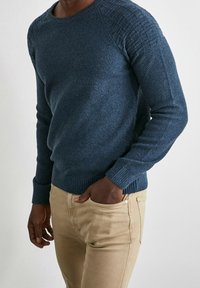 Mönstrad mörkblå stickad tröja med ribbade manschetter och nederkant, bärs med ljusbeiga byxor. Nära bild av vänster sida och arm.